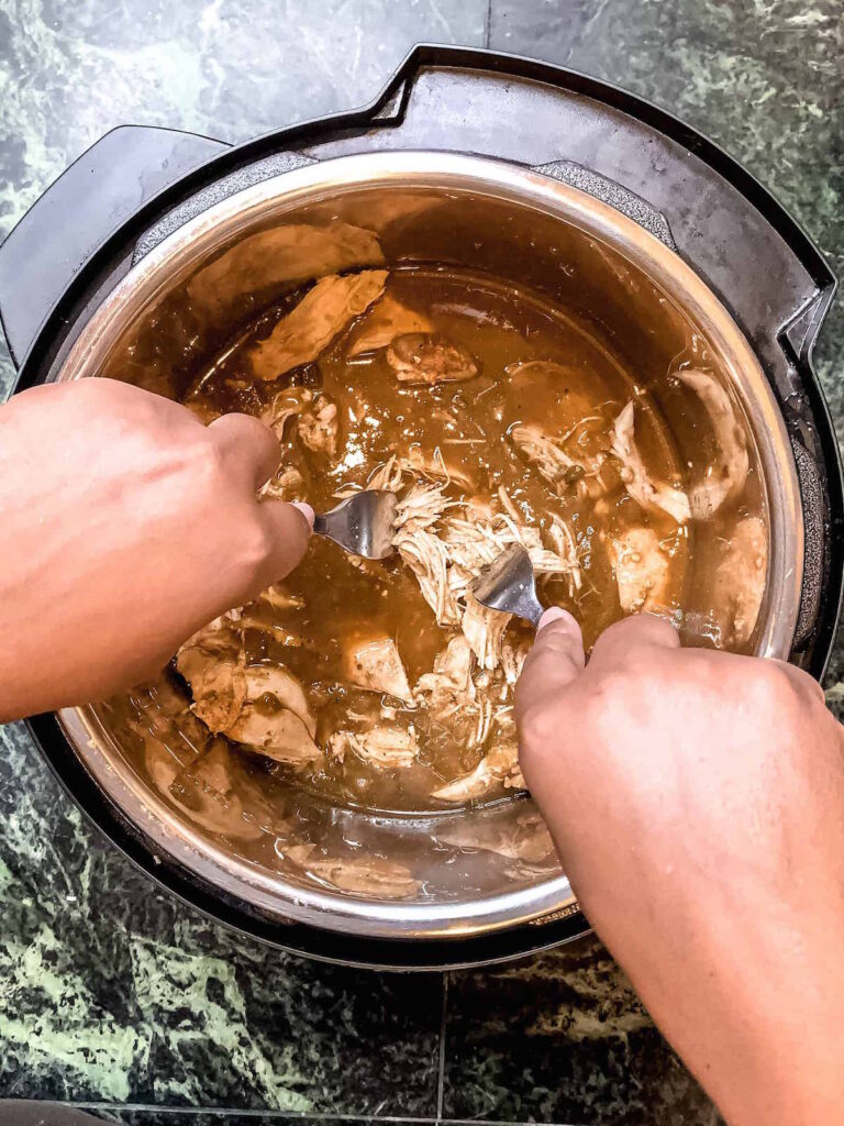 Brown hands holding two forks shredding chicken in salsa verde in the Instant Pot.