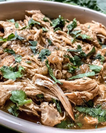 Shredded salsa verde chicken garnished with chopped cilantro in a ceramic dish.