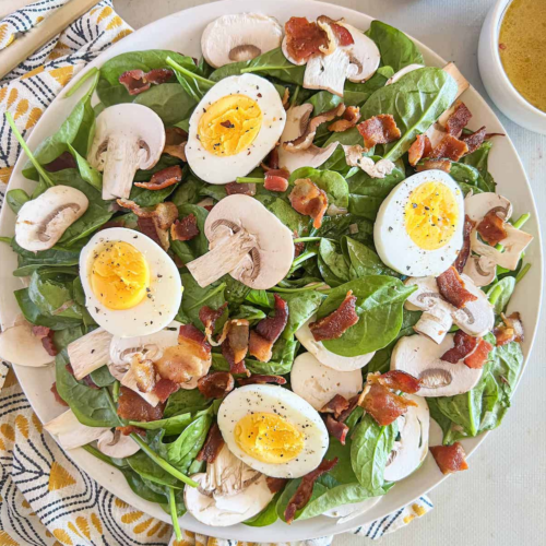 White plate of spinach salad topped with chopped bacon, sliced mushrooms and sliced hard-boiled eggs.