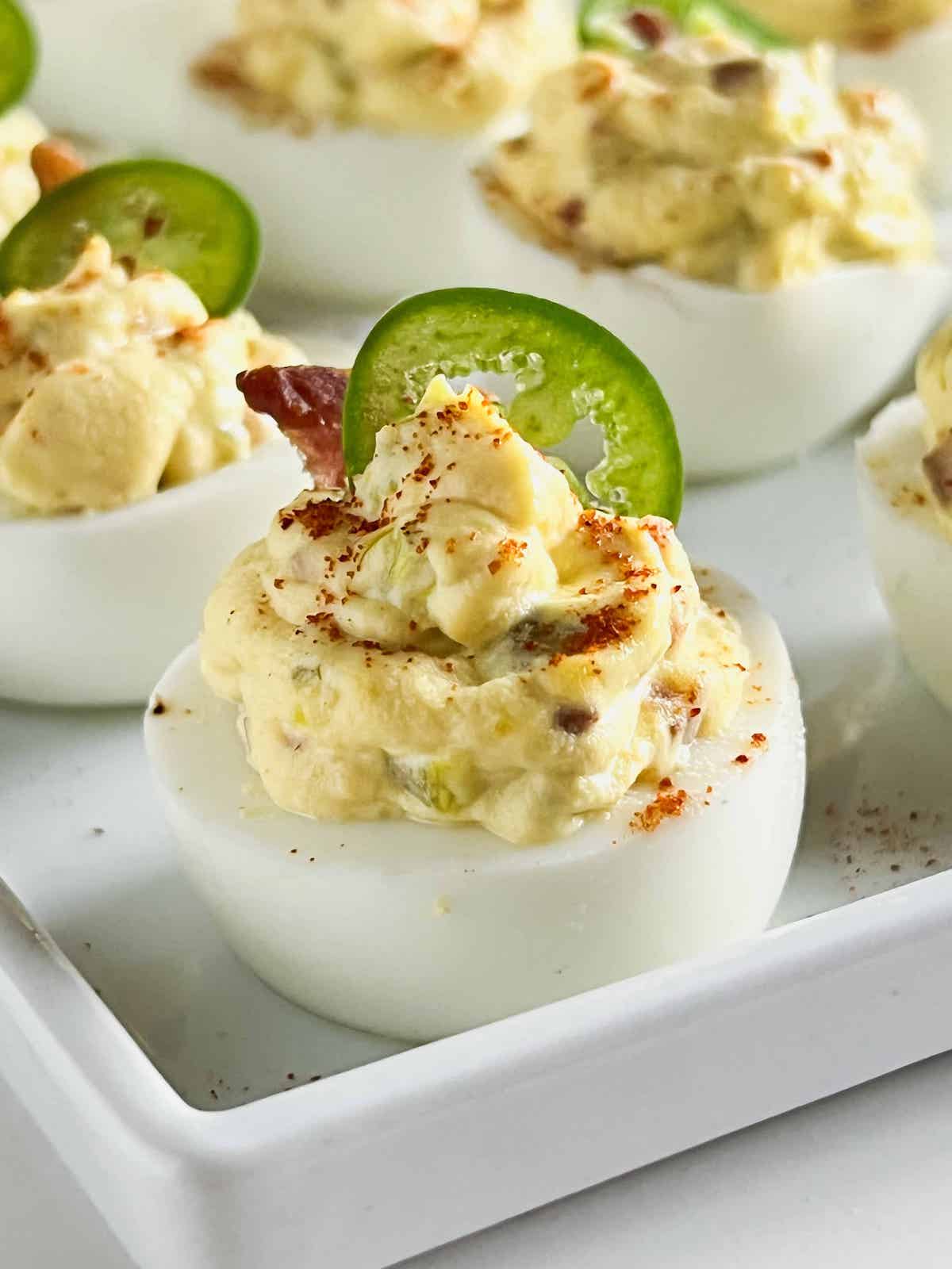 Close up of deviled egg on a white platter topped with a piece of bacon and a slice of jalapeno.