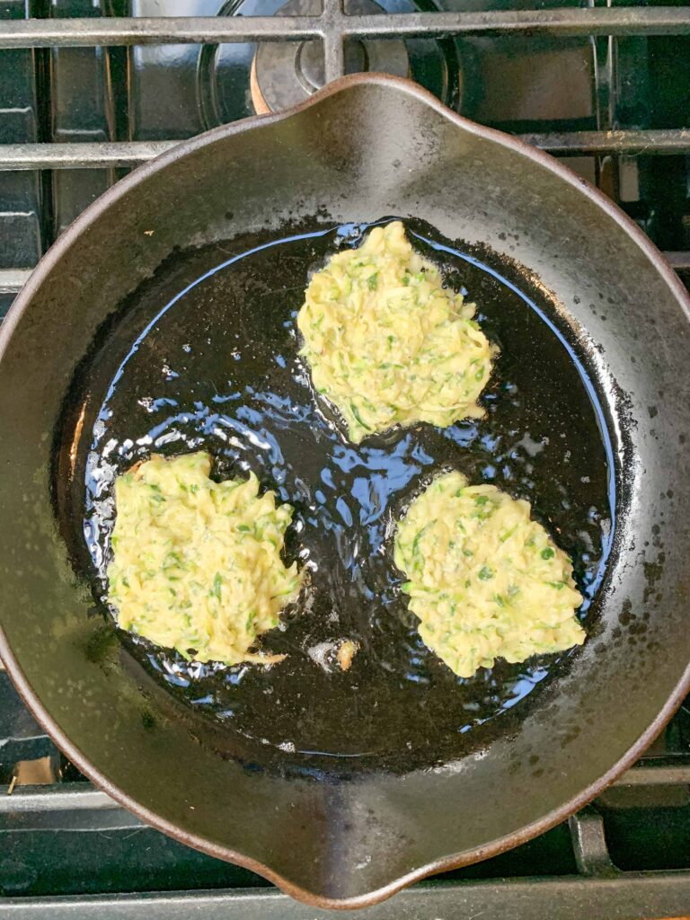 Zucchini fritters frying in a cast iron skillet.