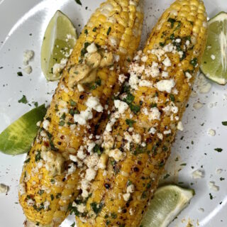 Corn on the cob topped with cotija cheese on a platter with lime wedges.