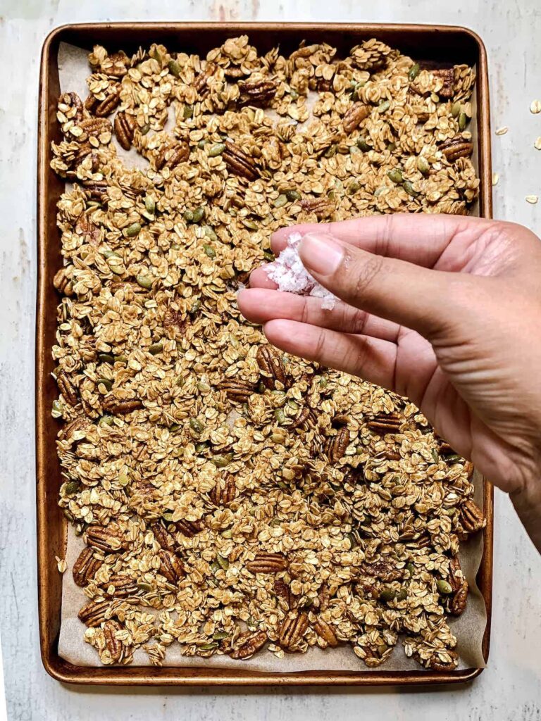 Baking sheet of granola with a brown hand sprinkling flaky salt.