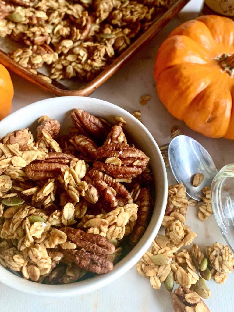 White bowl full of pumpkin pecan pie granola with a silver spoon next to it and a mini orange pumpkin and the corner of a baking sheet with more granola.