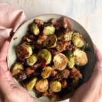 bowl of roasted brussels sprouts with bacon with hands setting on the table