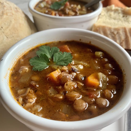 close up of hearty lentil soup in a whilte bowl with bread in the background