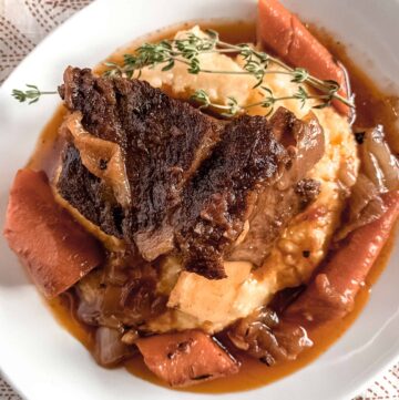 A white plate with creamy parmesan polenta topped with beer braised beef short ribs, carrots and a sprig of fresh thyme.