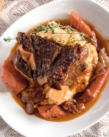 A white plate with creamy parmesan polenta topped with beer braised beef short ribs, carrots and a sprig of fresh thyme.
