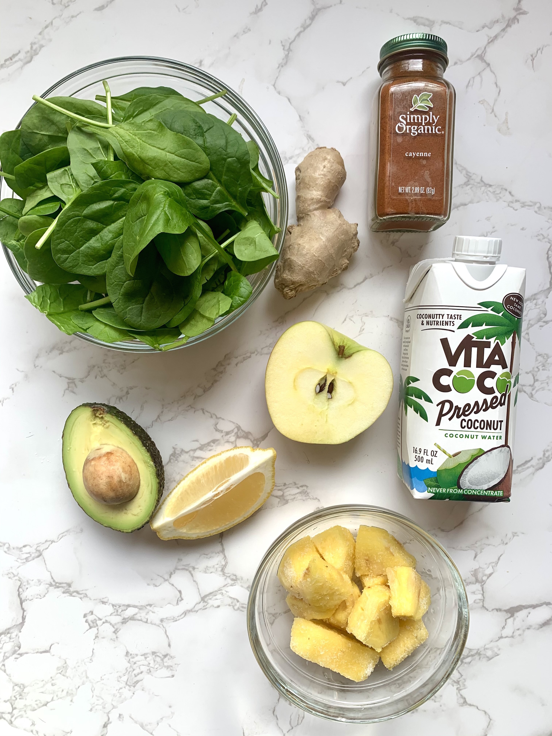 Ingredients laid out for green machine smoothie. Bowl of spinach leaves, half an avocado, half an apple, fresh ginger, bowl of pineapple chunks, carton of coconut water and bottle of cayenne pepper