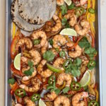 Sheet pan with shrimp, bell peppers, onions, cilantro leaves and lime wedges with charred blue corn tortillas