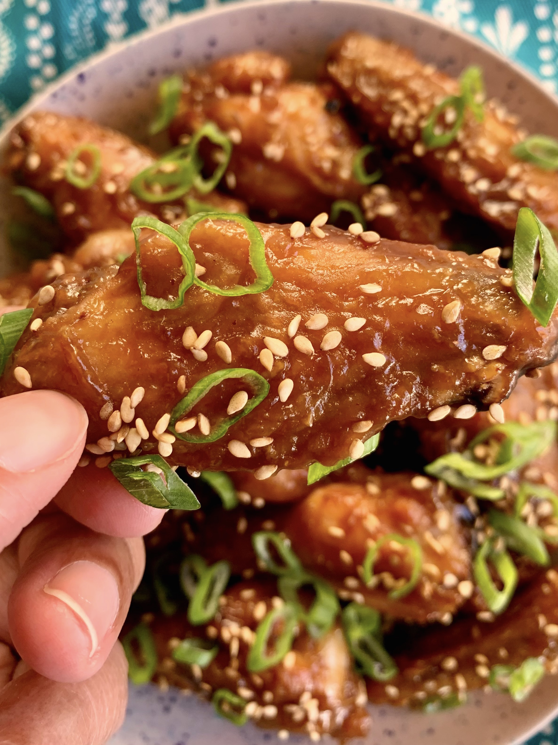 close up of hand holding wing flat garnished with sesame seeds and scallions. plate of wings in the background