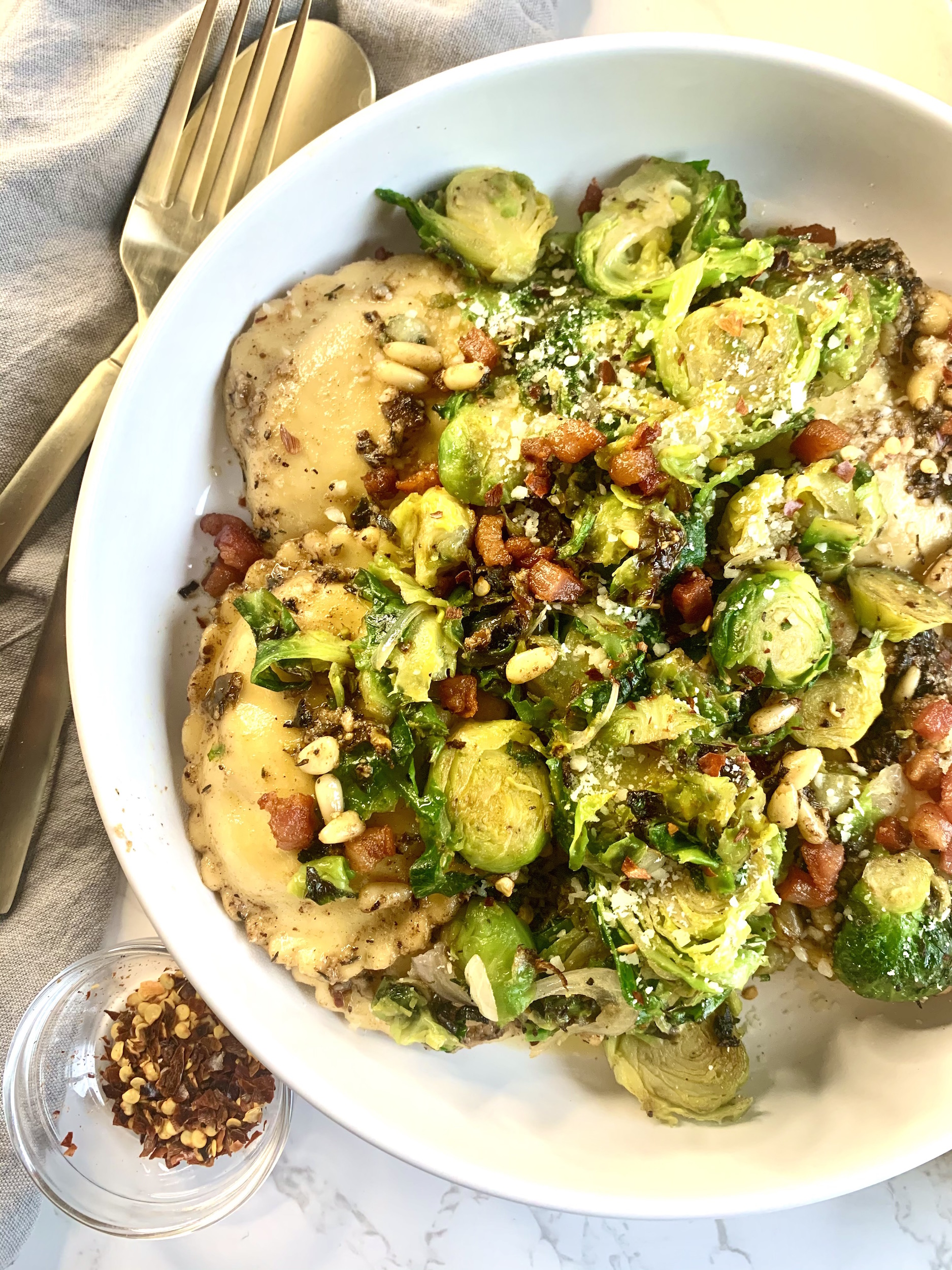 White plate with oval ravioli topped with shredded brussels sprouts and crispy pancetta.