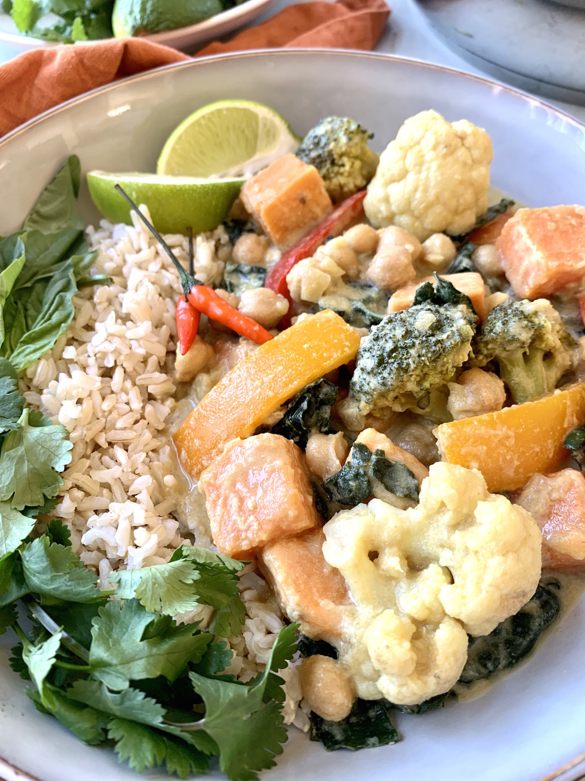 Close up of bowl of curry with broccoli, cauliflower, sweet potatoes, peppers and brown rice. garnished with herbs and lime wedges.