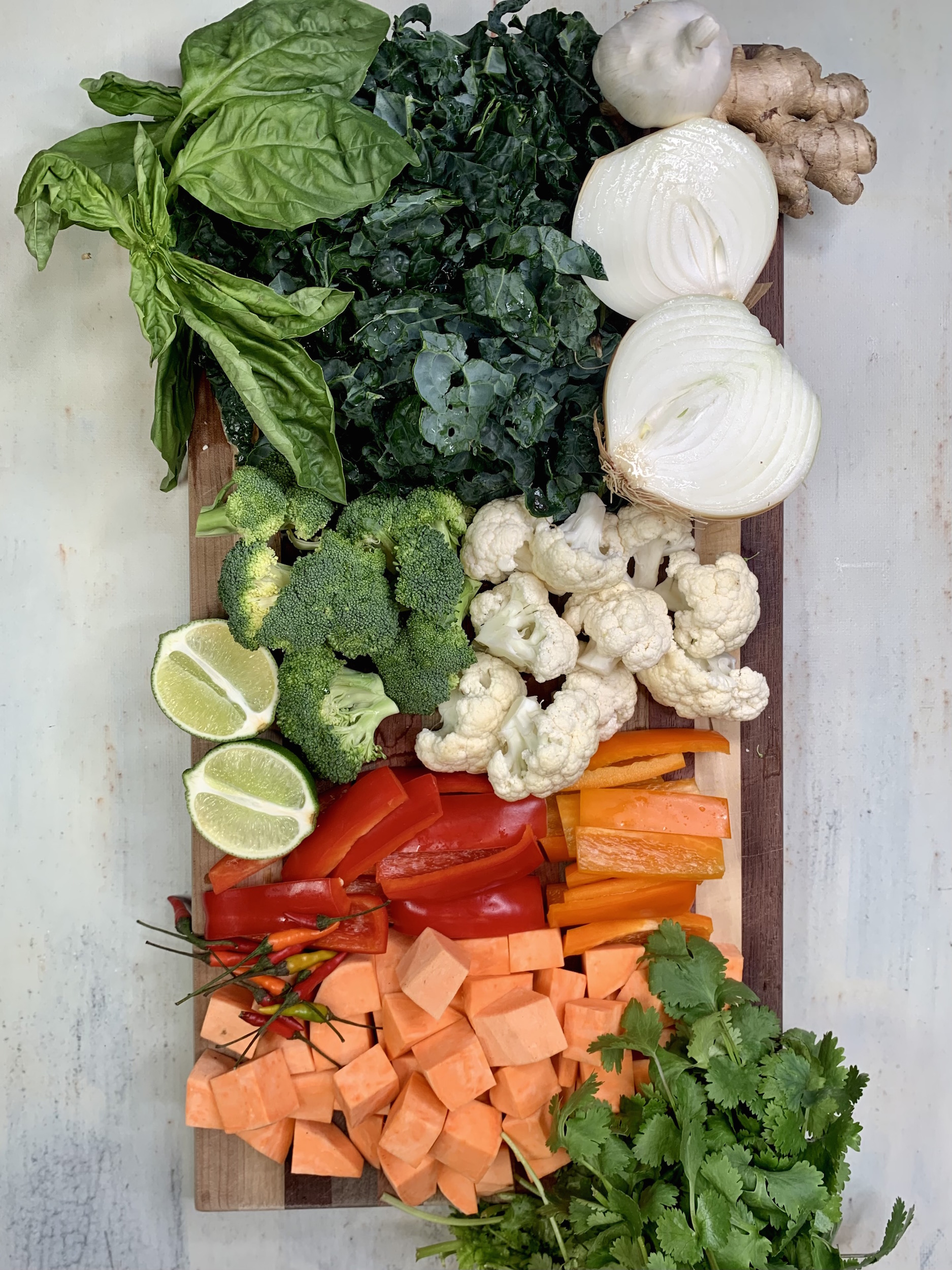 Onion, pepper, garlic, sweet potato, broccoli, cauliflower, basil, cilantro, ginger, lime and chili on a cutting board
