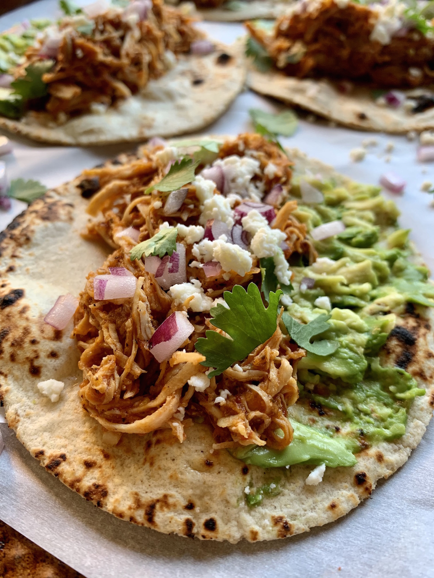 Close up of charred tortilla with chicken tinga, smashed avocado, diced onion, queso fresco and cilantro leaves