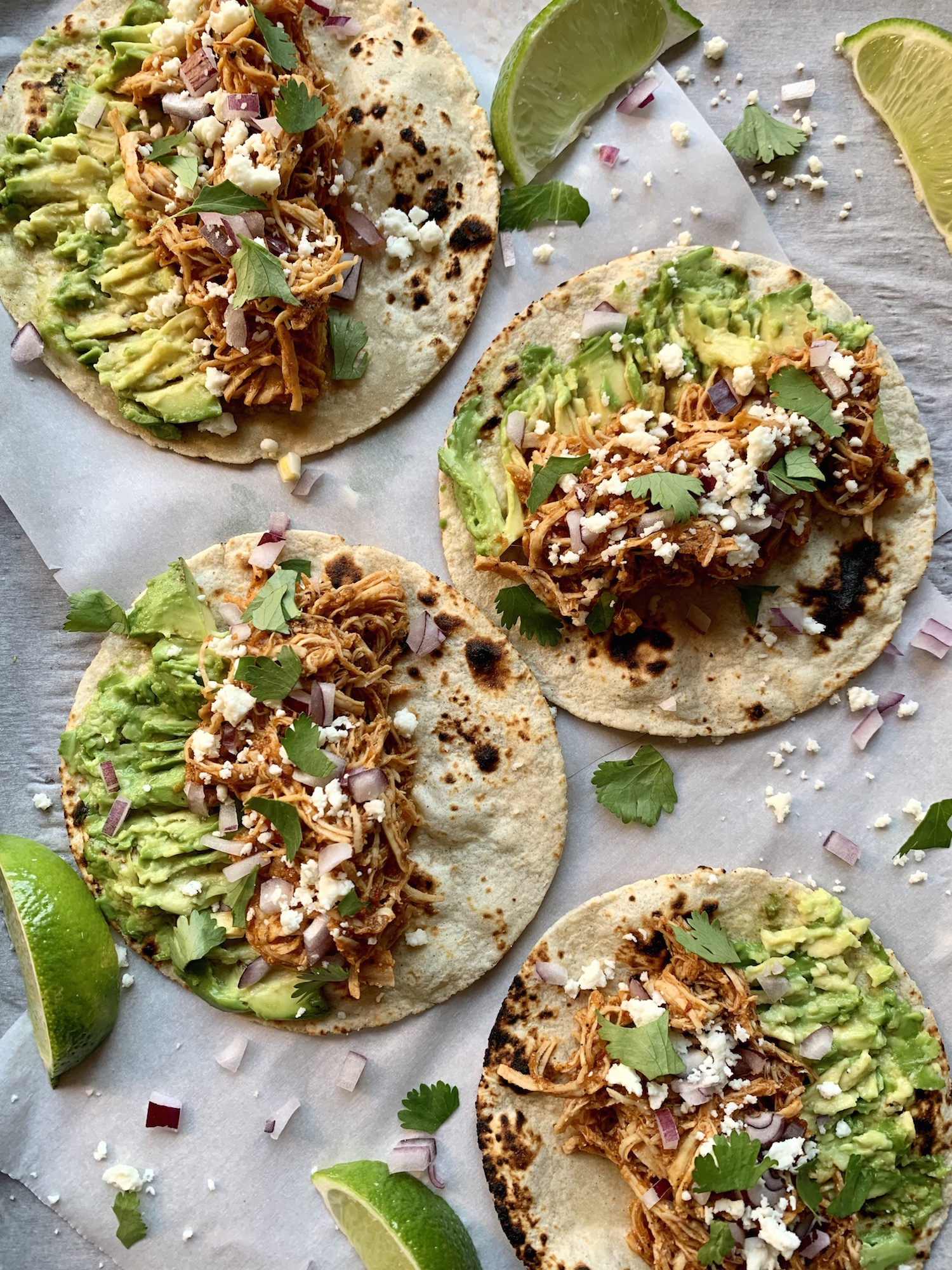 Soft tortillas open on parchment paper filled with chicken tinga, smashed avocado, queso fresco, diced onion and cilantro