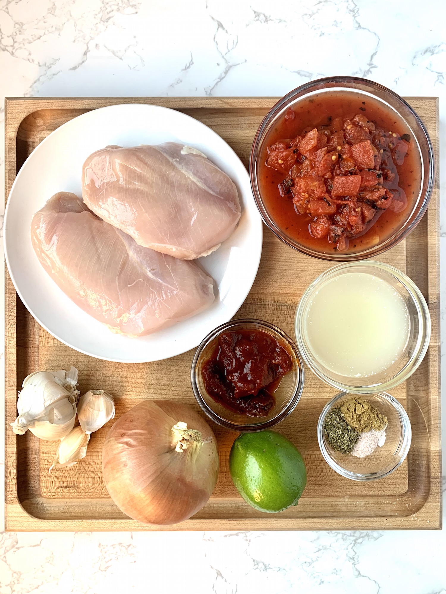 Chicken tinga ingredients in individual bowls on a wood cutting board - raw chicken breasts, fire roasted tomatoes, chicken broth, chipotle chilis in adobo, onion, garlic, lime and spices