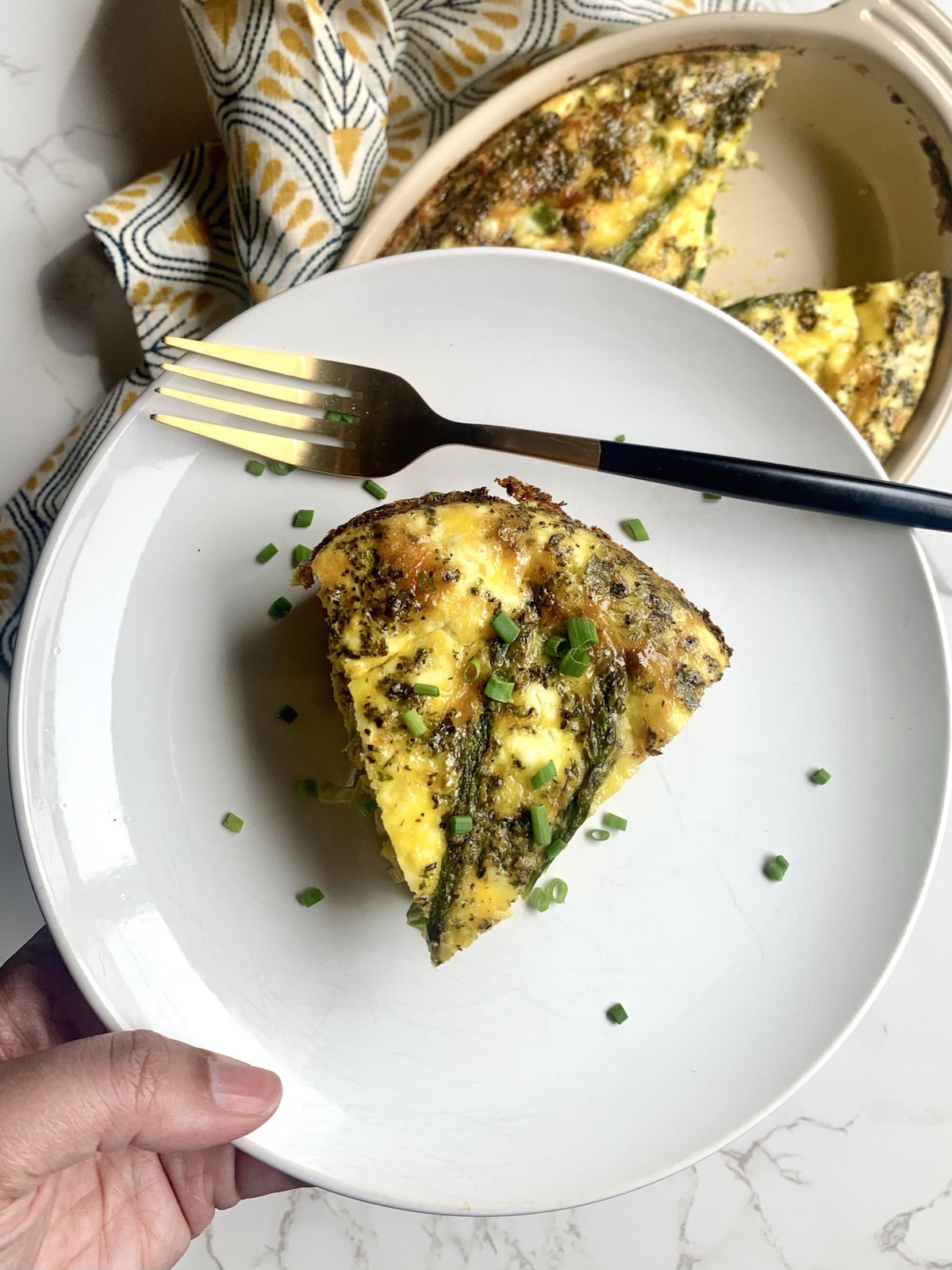 Hand holding a slice of baked asparagus, leek and goat cheese frittata on a white plate with a gold fork