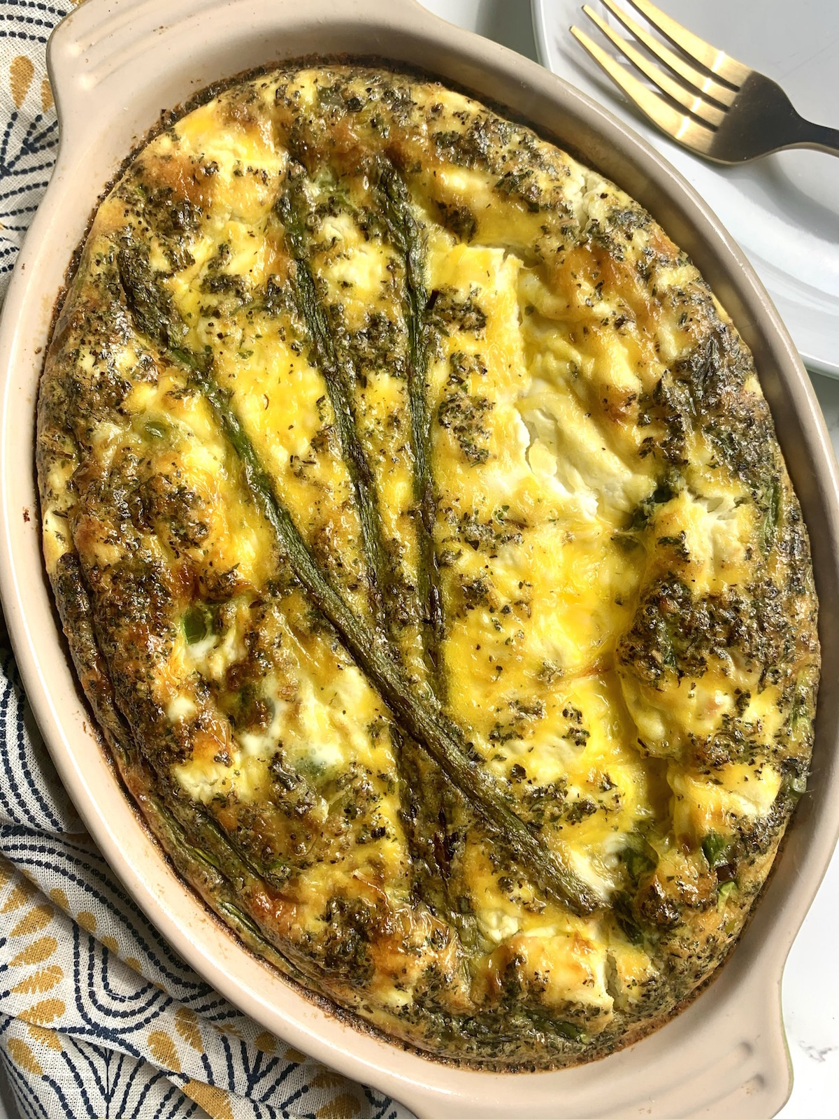 Baked asparagus, leek and goat cheese frittata in an oval baking dish with a yellow and gray napkin and white plate in the background