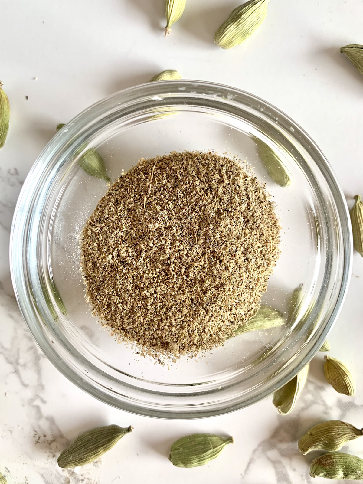 Small glass bowl of cardamom powder with whole pods in the background