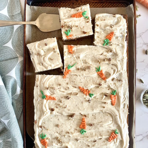 Cardamom Spiced Carrot Cake with Brown Butter Cream Cheese Frosting on parchment paper on a sheet pan with a silver cake server. cardamom pods sprinkled in the background