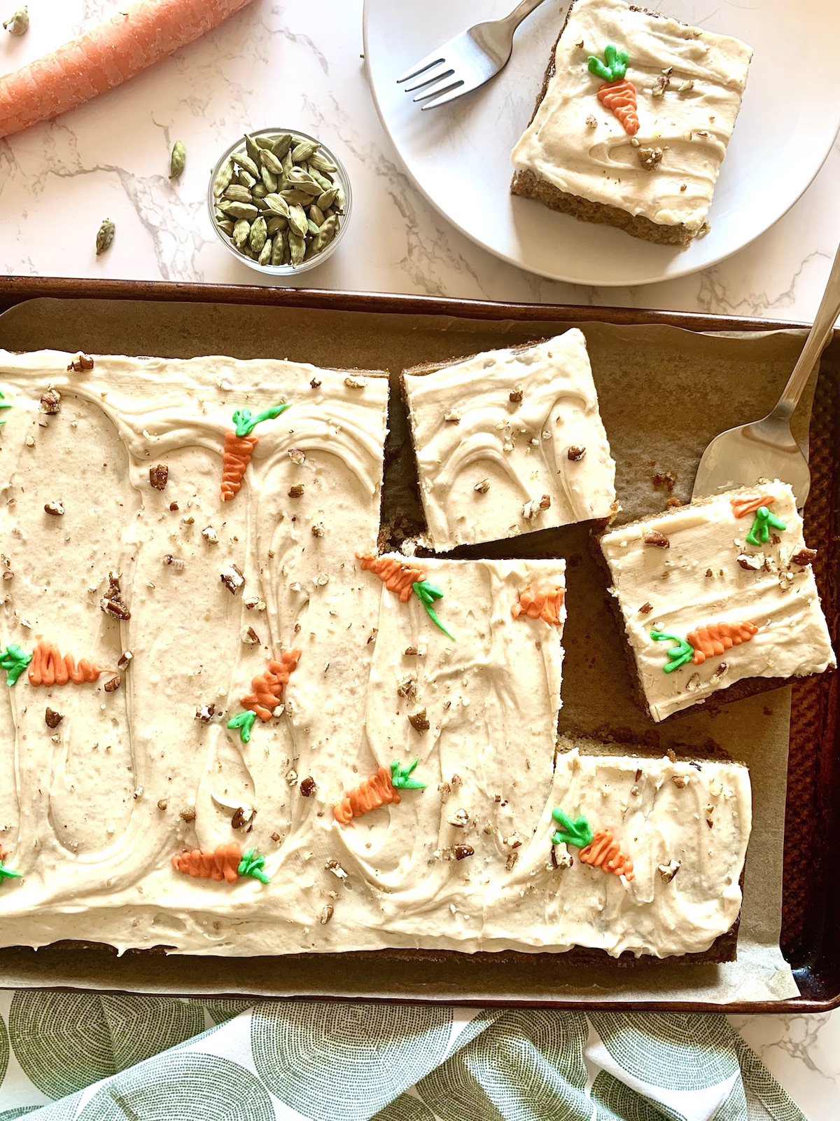 Full sheet of carrot cake sheet cake with brown butter cream cheese frosting with a few slices cut. one slice on a white plate and bowl of cardamom pods in the background