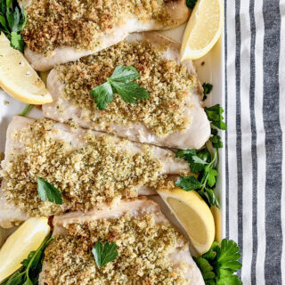 Overhead image of a platter of baked Panko-crusted white fish with parsley and lemon on a striped tablecloth.