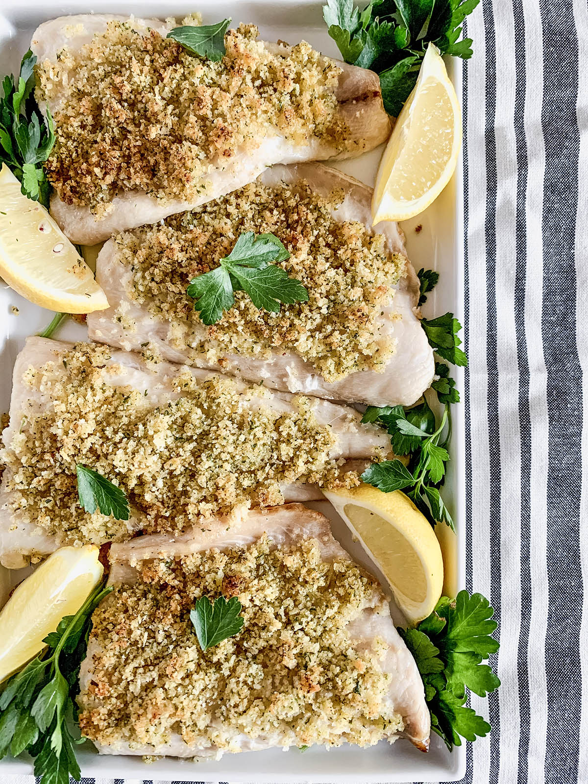 Overhead image of a platter of baked Panko-crusted white fish with parsley and lemon on a striped tablecloth.