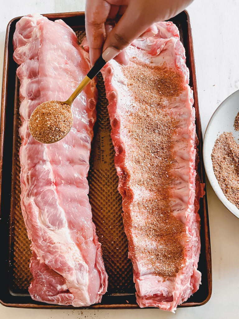 Two slabs of ribs on a baking sheet with a hand holding a gold spoon of dry rub