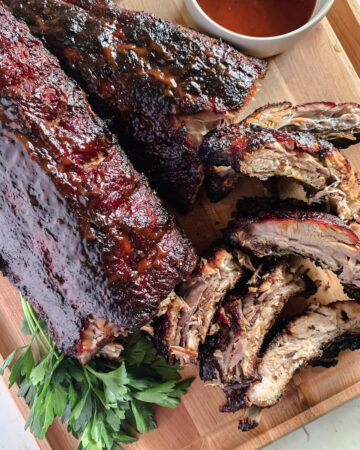 BBQ baby back ribs on a wooden platter with a small bowl of bbq sauce, garnished with parsley