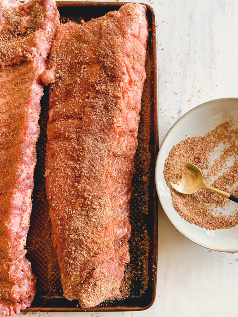 Dry rubbed slabs of ribs on a baking sheet next to a grey bowl with a gold spoon