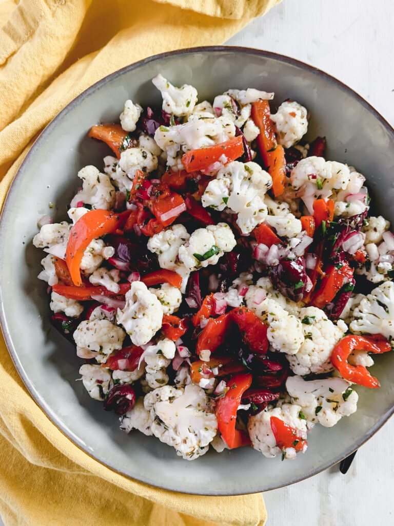 Marinated cauliflower salad with roasted peppers, kalamata olives and red onion in a grey bowl with a yellow napkin