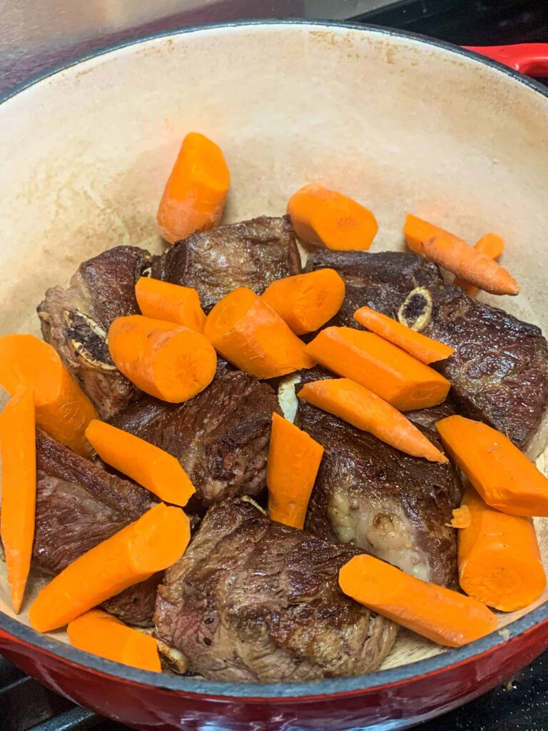 Seared beef short ribs transferred to dutch oven with raw carrot chunks on top