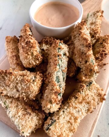 Crispy air fryer garlic parmesan zucchini fries on a wood board with a white bowl of dipping sauce.
