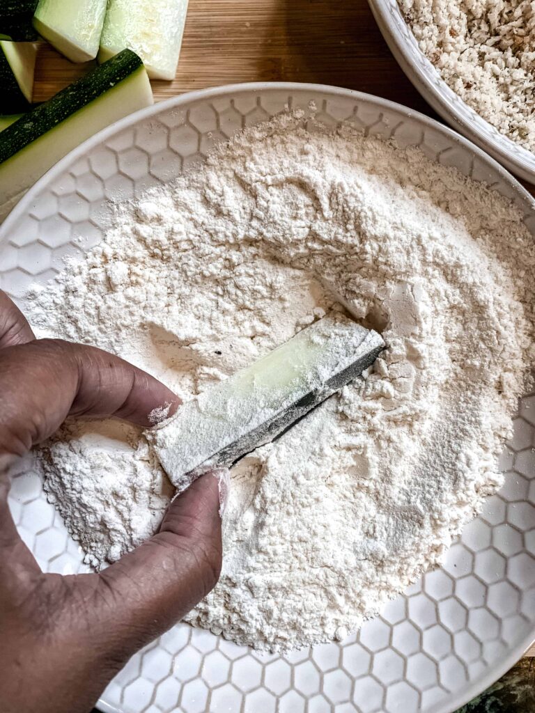 Hand holding a zucchini stick dredging in flour mixture.
