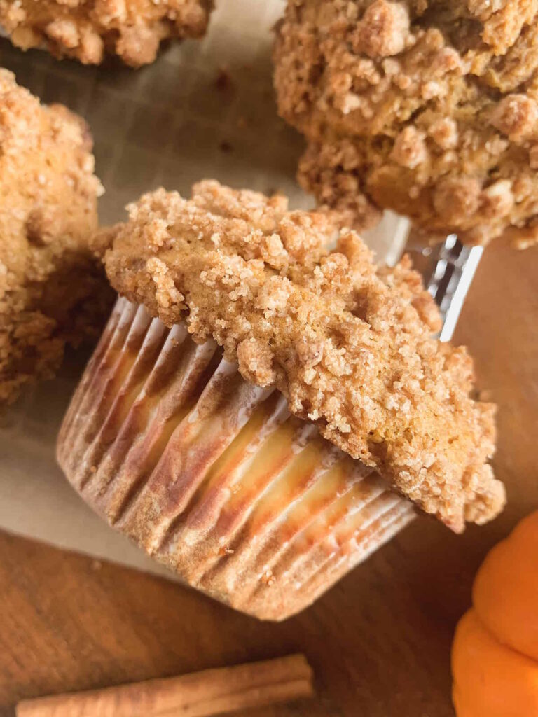 Close up of spiced pumpkin cream cheese muffin with cinnamon streusel topping on parchment paper with an orange pumpkin and cinnamon stick.