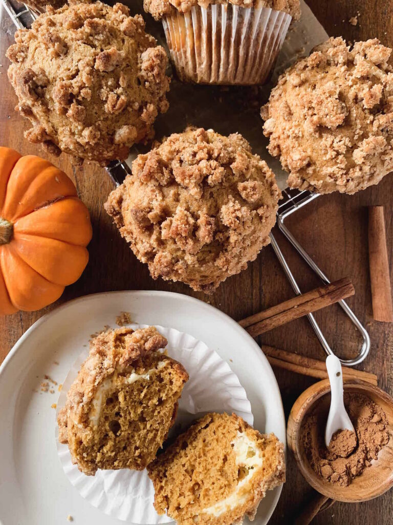 Spiced pumpkin cream cheese muffins with streusel topping on a wire rack with a split muffin on a white plate and cinnamon sticks, small bowl of pumpkin spice and mini orange pumpkin.