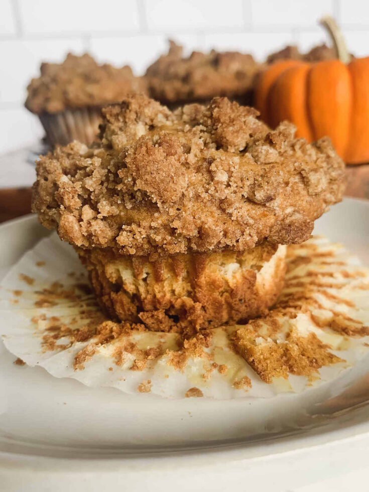 Spiced pumpkin cream cheese muffin with cinnamon streusel topping on a peeled muffin wrapper with muffins and a mini orange pumpkin in the background.