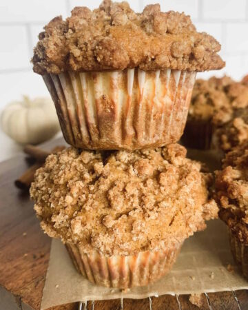 Two spiced pumpkin muffins stacked on top of each other with more muffins and a small white pumpkin in the background.
