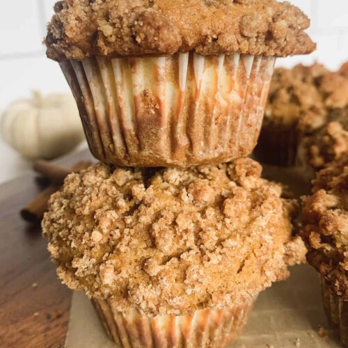 Two spiced pumpkin muffins stacked on top of each other with more muffins and a small white pumpkin in the background.