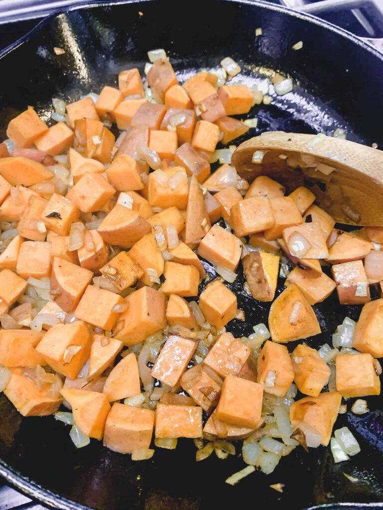 Sweet potato cubes sauteeing with diced onions in a cast iron skillet with a wooden spoon. 