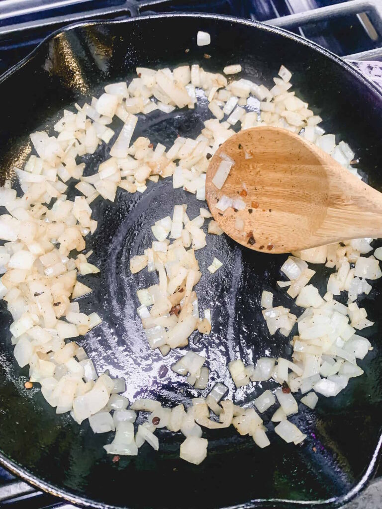 Diced onions sauteeing in a cast iron skillet with a wooden spoon.