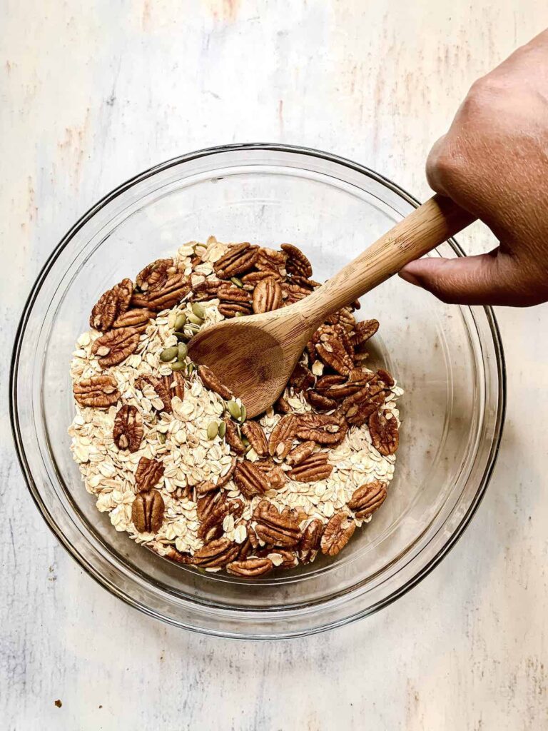 Brown hand with a wooden spoon stirring together oats, pepitas and pecans in a large glass bowl.