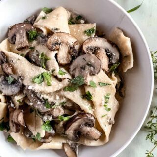 White bowl full of pappardelle with mushrooms and creamy garlic parmesan sauce with fresh herbs in the background.