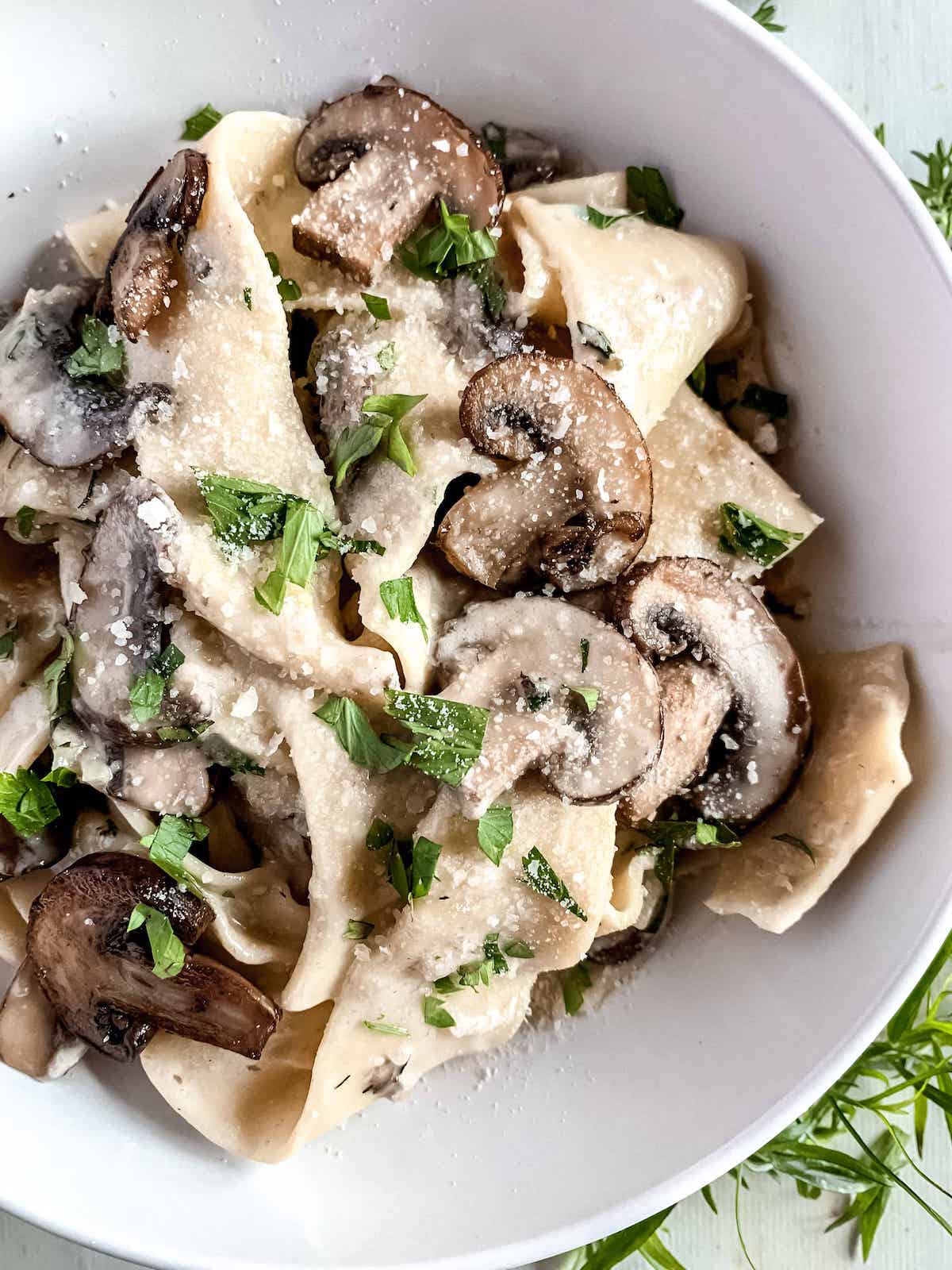 Pappardelle pasta with mushrooms in creamy garlic parmesan sauce topped with fresh herbs in a white bowl.