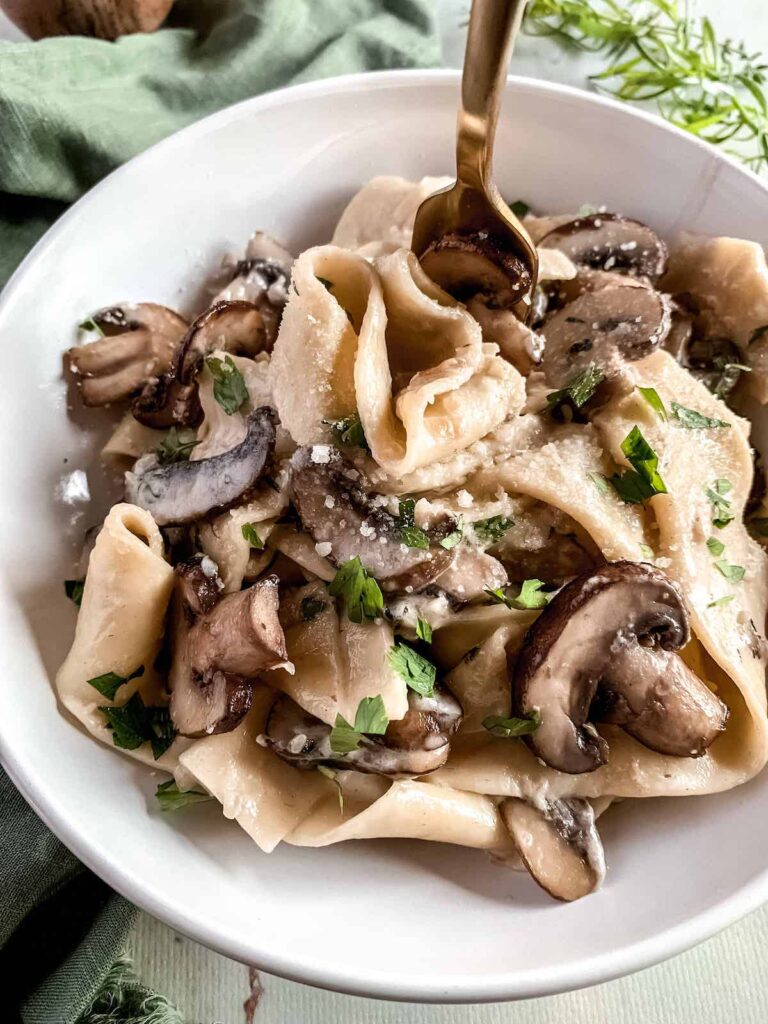 White bowl with pappardelle pasta and mushrooms in creamy garlic parmesan sauce with a gold fork.