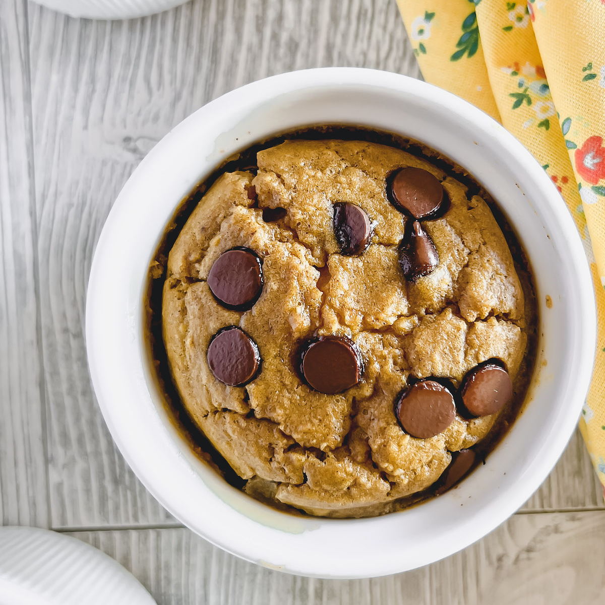 White ramekin with pumpkin blended baked oats topped with chocolate chips and a yellow floral napkin in the background.