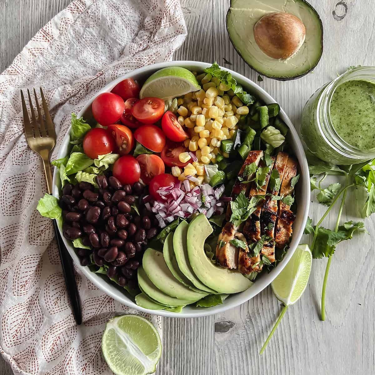 White dish with southwest salad topped with honey chipotle chicken on a table with a white patterned napkin, lime wedge, cilantro sprigs and a jar of dressing.