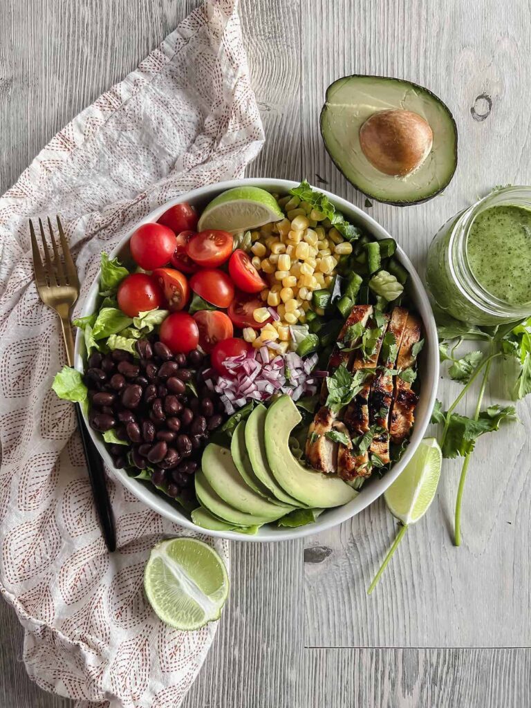 Overhead view of Southwest Salad with Honey Chipotle Chicken in a white bowl. Gold pattern napkin, gold fork, half an avocado, lime wedges, cilantro and jar of cilantro lime dressing on the table with salad.