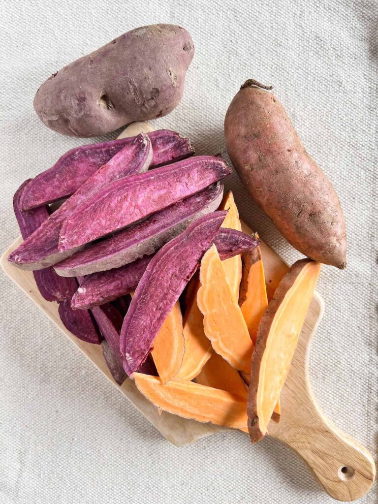 Small wood cutting board with cut wedges of orange and purple sweet potatoes and whole sweet potatoes next to it.
